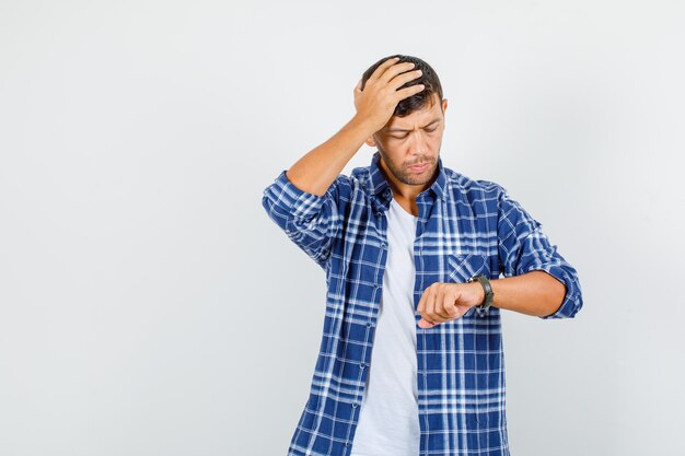 Young man in shirt looking at watch with hand on head and looking worried , front view.