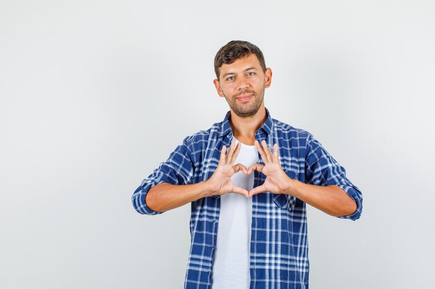 Young man in shirt making heart shape gesture and smiling , front view.