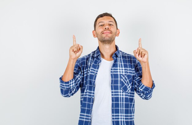 Young man in shirt pointing up fingers with closed eyes and looking confident , front view.