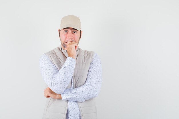 Young man in shirt,sleeveless jacket,cap holding fist on his dropped jaw and looking excited , front view. space for text