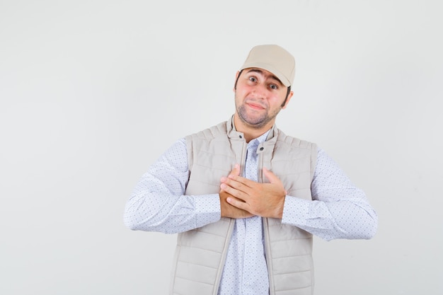 Giovane uomo in camicia, giacca senza maniche, berretto tenendosi per mano sul petto e guardando grato, vista frontale.