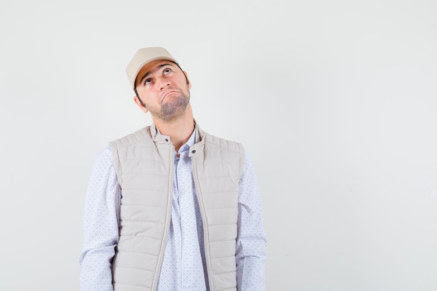 Young man in shirt,sleeveless jacket,cap looking up and looking pensive , front view.