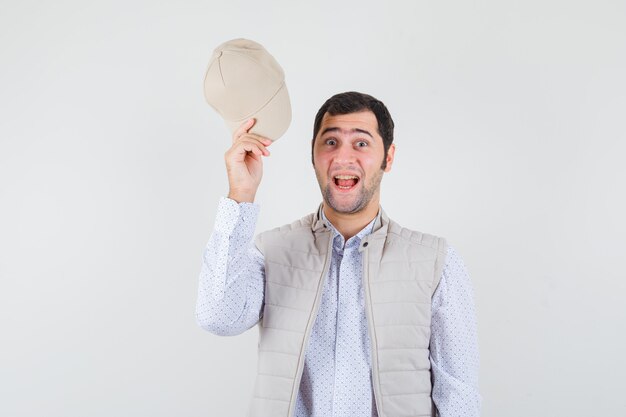 Young man in shirt,sleeveless jacket holding cap while smiling and looking happy , front view.