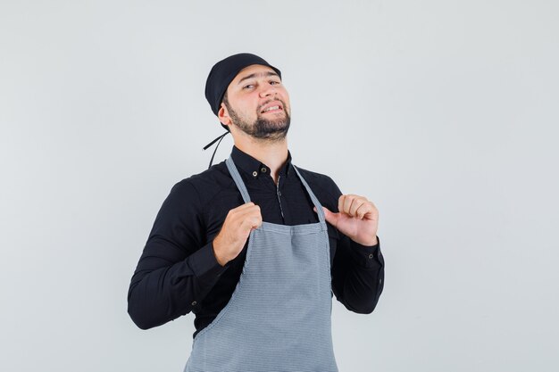 Young man in shirt standing with hands touching apron and looking confident , front view.