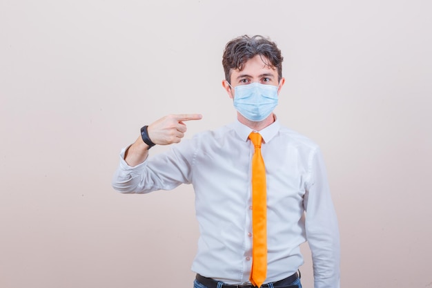 Young man in shirt, tie, jeans pointing at his medical mask