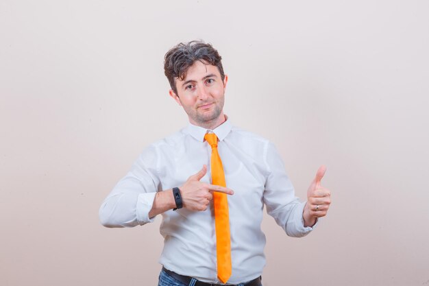 Young man in shirt, tie, jeans pointing at his thumb up and looking confident