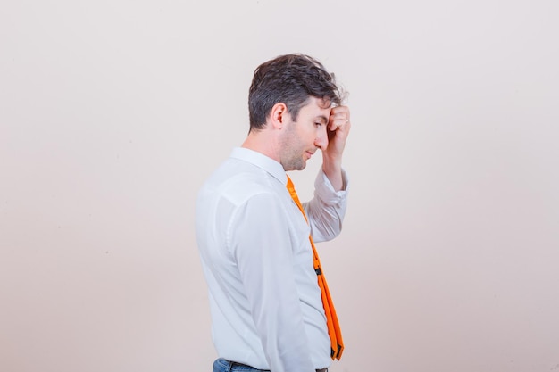 Young man in shirt, tie, jeans rubbing forehead and looking pensive .