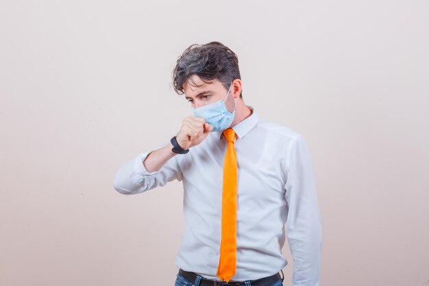 Young man in shirt, tie, mask, jeans suffering from cough and looking unwell