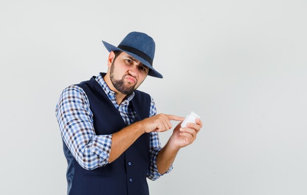 Young man in shirt, vest, hat pointing at bottle of pills and looking pensive , front view.