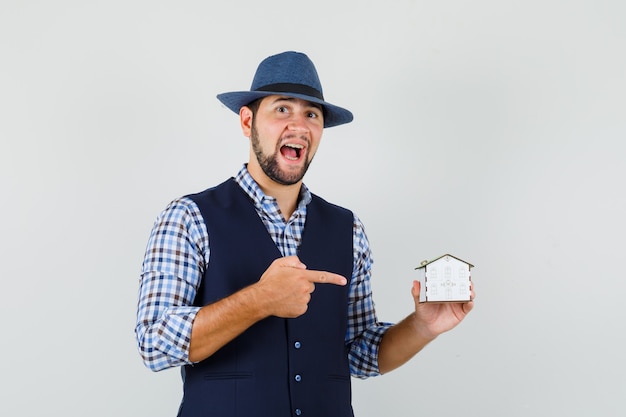 Young man in shirt, vest, hat pointing at house model and looking confident , front view.