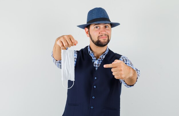 Young man in shirt, vest, hat pointing at medical mask and looking optimistic , front view.