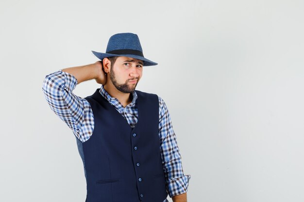 Young man in shirt, vest, hat posing with hand on neck and looking elegant , front view.