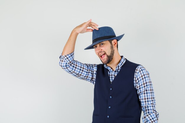 Young man in shirt, vest holding fingers on his hat and looking confident , front view.