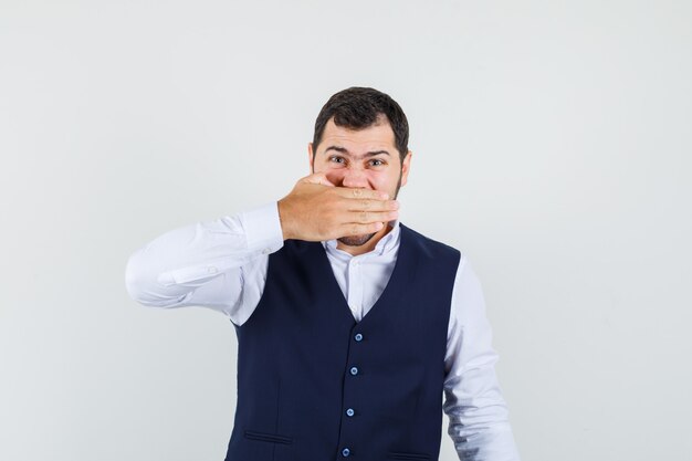 Young man in shirt, vest holding hand over mouth and looking irritated