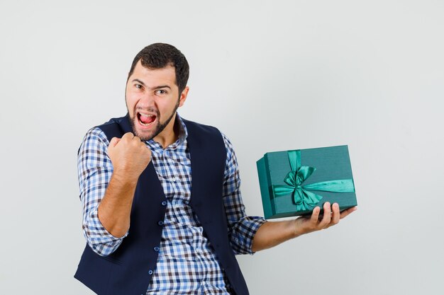 Young man in shirt, vest holding present box, showing winner gesture and looking happy , front view.