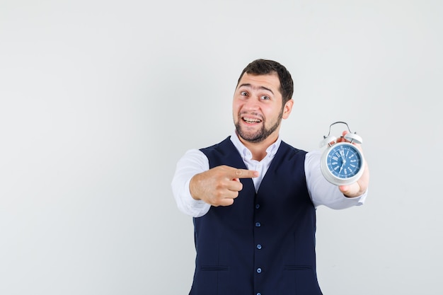 Young man in shirt, vest pointing at alarm clock and looking optimistic