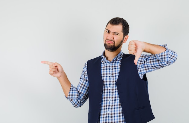 Young man in shirt, vest showing thumb down, pointing to the side and looking disappointed , front view.