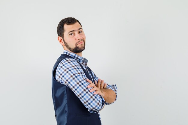 Young man in shirt, vest standing with crossed arms and looking confident .