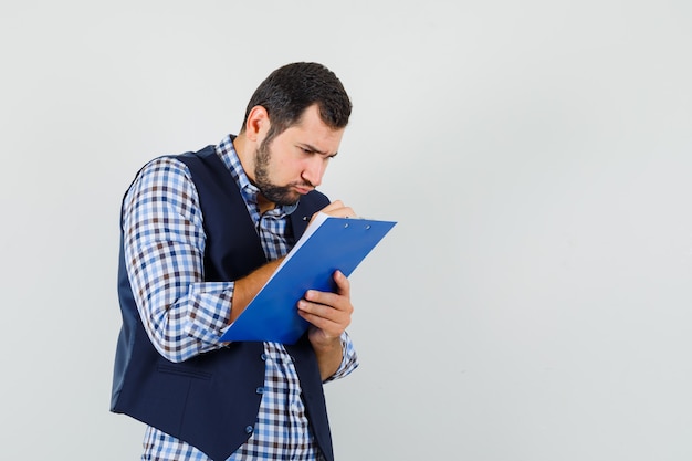 Young man in shirt, vest taking notes on clipboard and looking busy , front view.