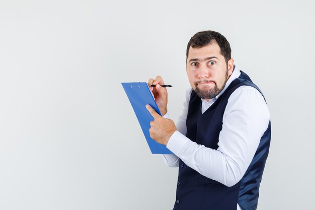 Young man in shirt, vest taking notes on clipboard and looking perplexed, front view.