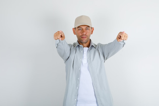 Young man in shirt with cap pointing fingers to camera and looking confident