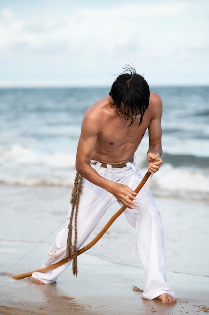 Молодой человек без рубашки на пляже с деревянным луком готовится заниматься капоэйрой