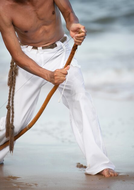 カポエイラを練習する準備をしている木製の弓とビーチで上半身裸の若い男