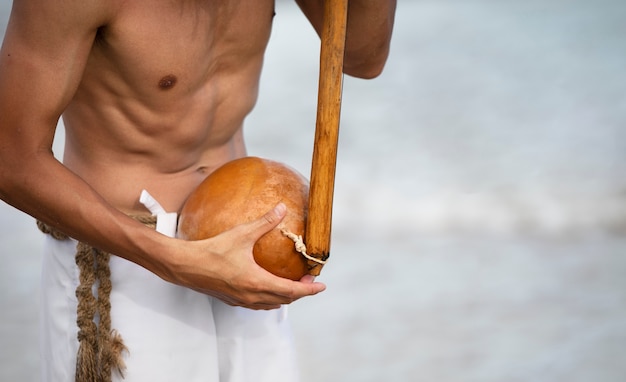 Young man shirtless on the beach with wooden bow preparing to practice capoeira