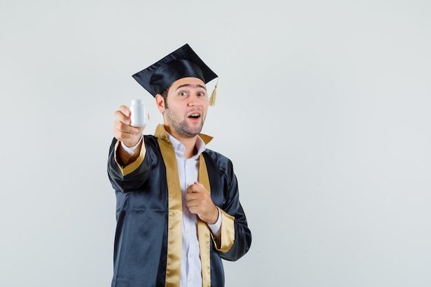 Young man showing bottle of pills in graduate uniform and looking happy , front view.