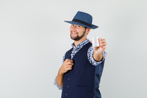 Foto gratuita giovane che mostra una bottiglia di pillole in camicia, gilet, cappello e sembra allegro