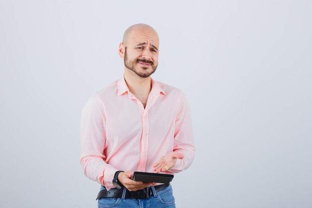 Young man showing calculator in pink shirt,jeans and looking displeased. front view.