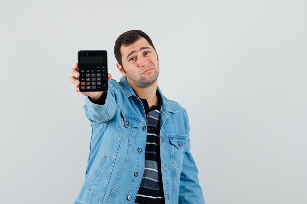 Young man showing calculator in t-shirt, jacket and looking desperate , front view.