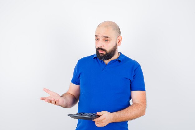 Young man showing calculator while talking in blue shirt and looking confused , front view.