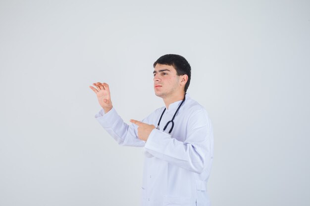 Young man showing claw gesture, pointing to the left side in doctor uniform and looking cheerful