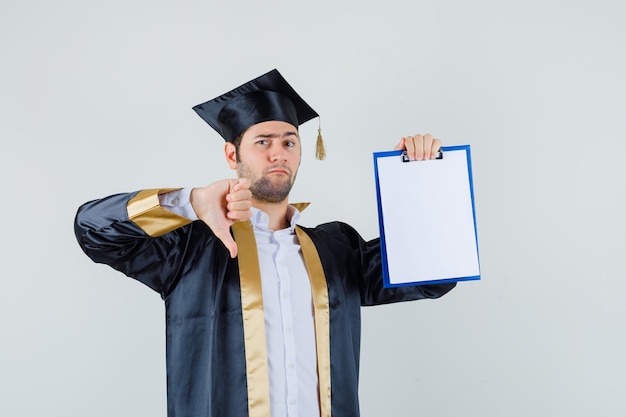 Free photo young man showing clipboard with thumb down in graduate uniform and looking gloomy , front view.