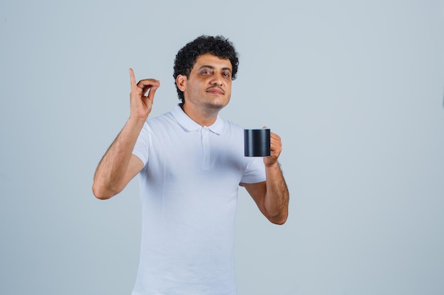 Young man showing delicious gesture and holding cup of tea in white t-shirt and jeans and looking happy , front view.