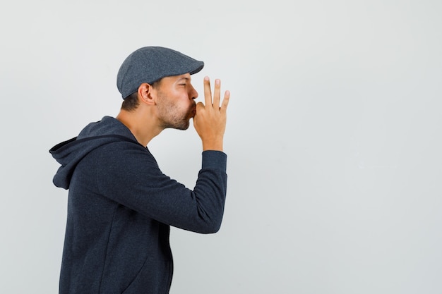 Young man showing delicious gesture in t-shirt, jacket, cap and looking delighted. .