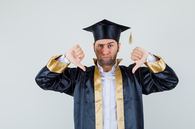 Young man showing double thumbs down in graduate uniform and looking serious. front view.