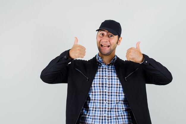 Young man showing double thumbs up in shirt, jacket, cap and looking glad , front view.