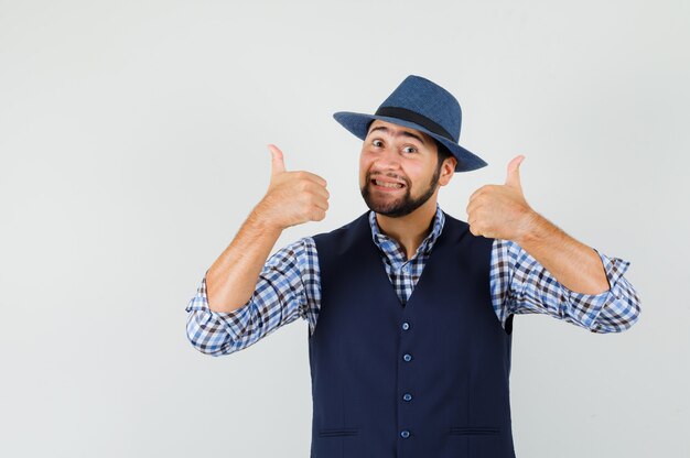 Young man showing double thumbs up in shirt, vest, hat and looking cheery.