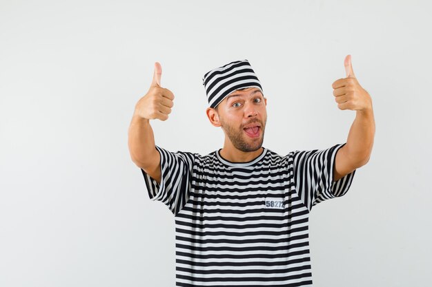 Young man showing double thumbs up in striped t-shirt, hat and looking jolly.