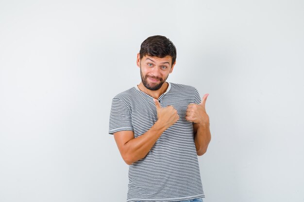Молодой человек показывает двойные пальцы вверх в полосатой футболке и выглядит веселым, вид спереди.