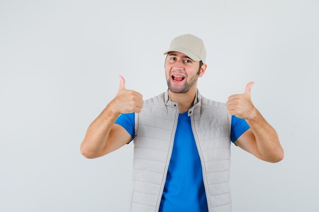 Young man showing double thumbs up in t-shirt, jacket, cap and looking happy. front view.