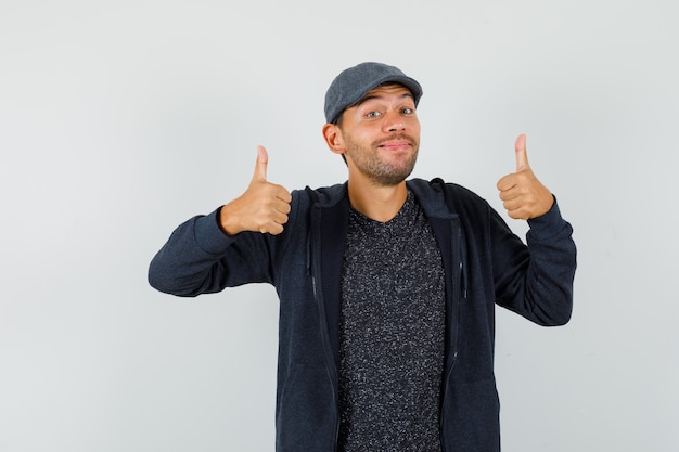 Young man showing double thumbs up in t-shirt, jacket, cap and looking merry