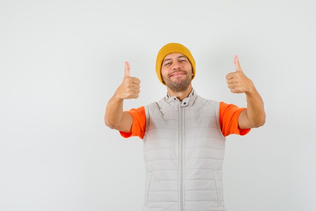 Young man showing double thumbs up in t-shirt, jacket, hat and looking cheerful.
