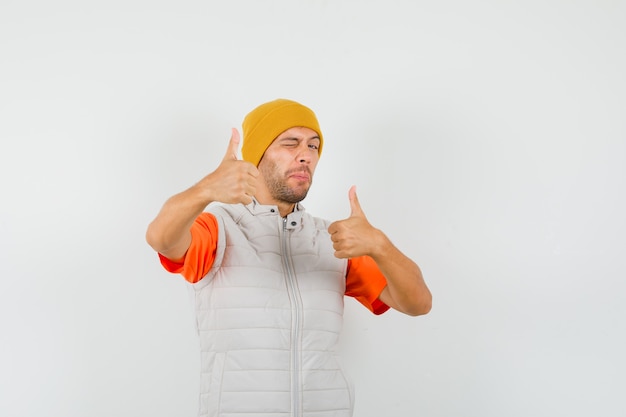 Young man showing double thumbs up, winking eye in t-shirt, jacket, hat