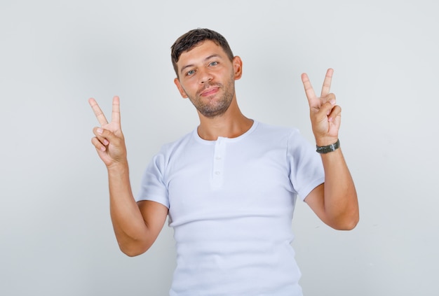 Молодой человек показывает пальцы, делает знак победы в белой футболке и выглядит счастливым, вид спереди