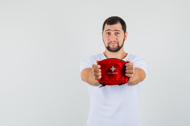 Young man showing first aid kit in white t-shirt and looking focused. front view. space for text