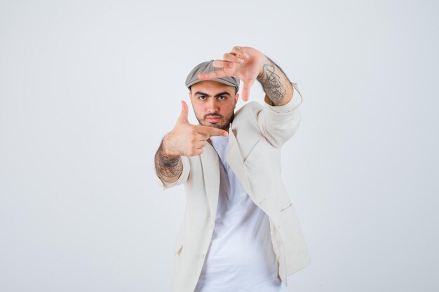 Young man showing frame gesture in white t-shirt, jacket and gray cap and looking optimistic. front view.