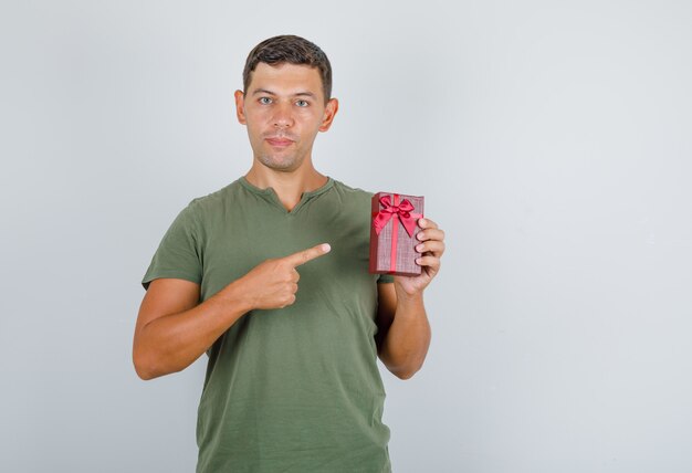 Young man showing gift box with finger in army green t-shirt front view.